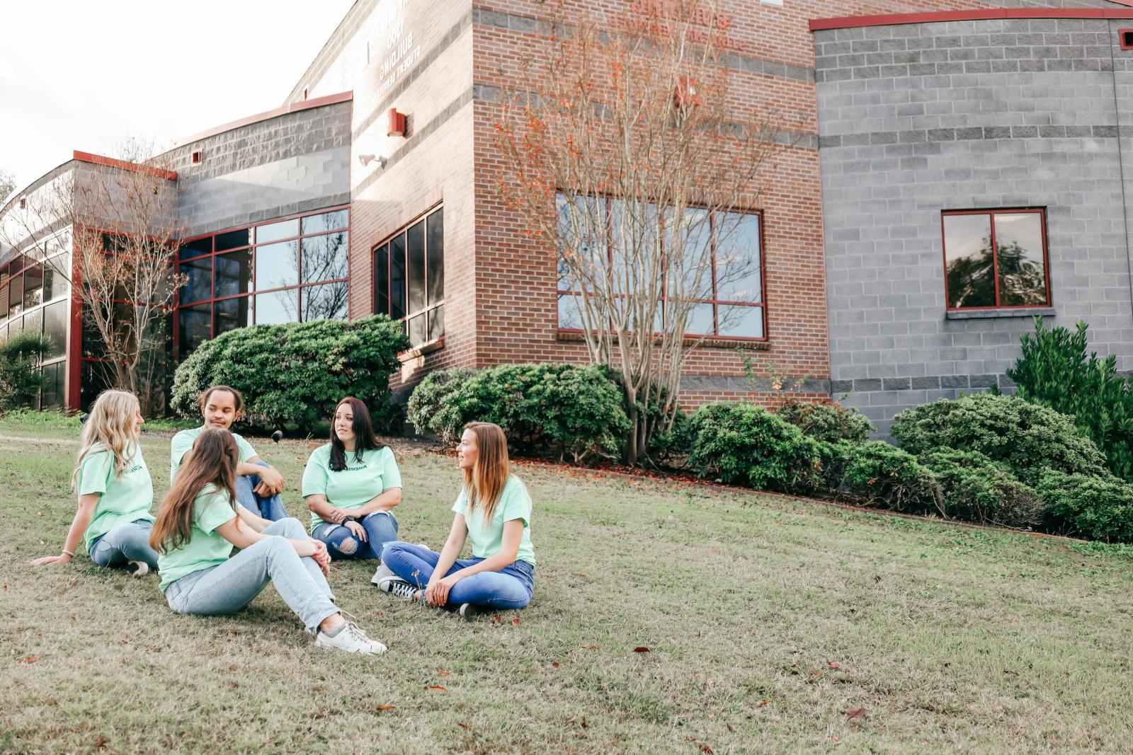 A group of students all wearing mint green Augusta Technical College shirts sit on the lawn to the left of the 1300 bldg chatting.